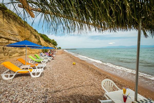 Strand des Pavlina Beach Hotels mit vielen Liegen, Sonnenschirmen und einem Tisch mit Getränken.