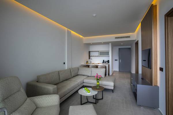 Wohnzimmer im Cavo Maris Beach Hotel mit L-förmiger Couch, Tisch, Fernseher und Küche im Hintergrund.