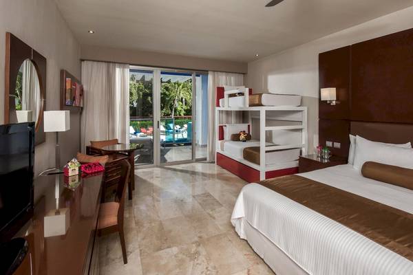 Innenansicht der Suite im Grand Riviera Princess Hotel mit Doppelbett, Etagenbett und Blick auf den Pool.