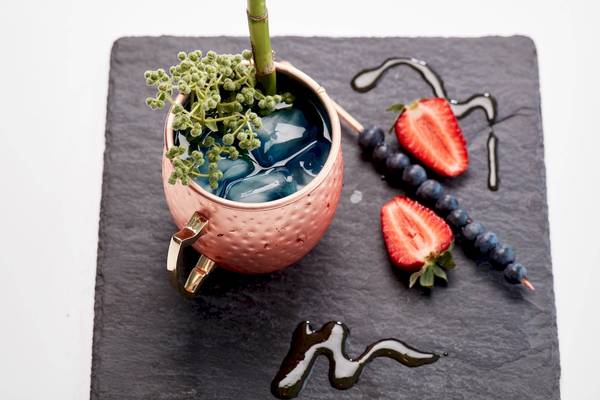 Ein Cocktail mit blauem Getränk, Eiswürfeln, Pfefferminz und Beeren auf einem grauen Tablett.