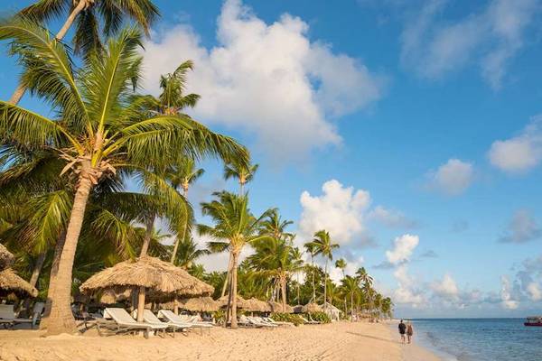 Strand des Catalonia Royal Bavaro mit Palmen, Sonnenliegen und zwei Personen, die am Wasser entlanggehen