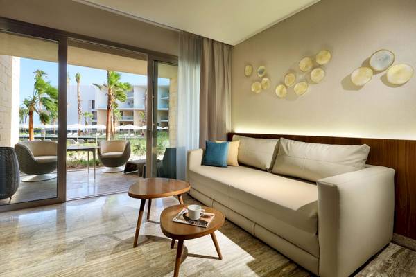 Wohnbereich im Grand Palladium Costa Mujeres Resort & Spa mit einer Couch, zwei Tischen und einer Aussicht auf Palmen und Gebäude.