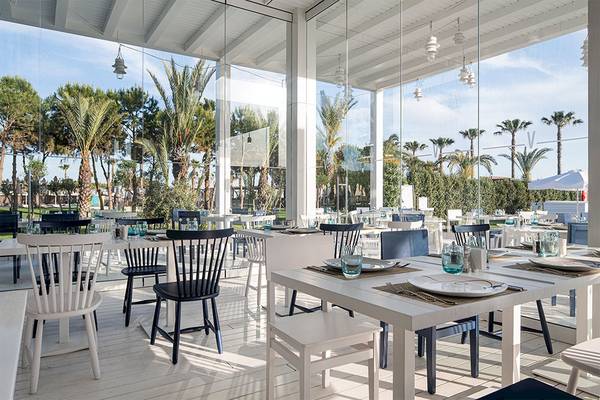 Innenansicht des Restaurants im Voyage Belek Golf & Spa Hotel mit weißen Tischen und Stühlen und Glaswänden.