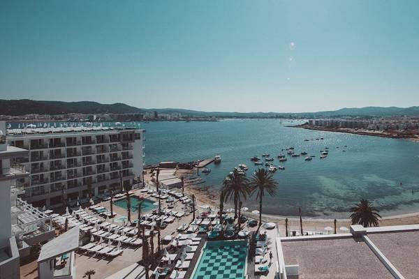 Aussicht von Amare Beach Hotel auf den Strand in Ibiza mit mehreren Gebäuden, Pools und vielen Booten im Wasser.