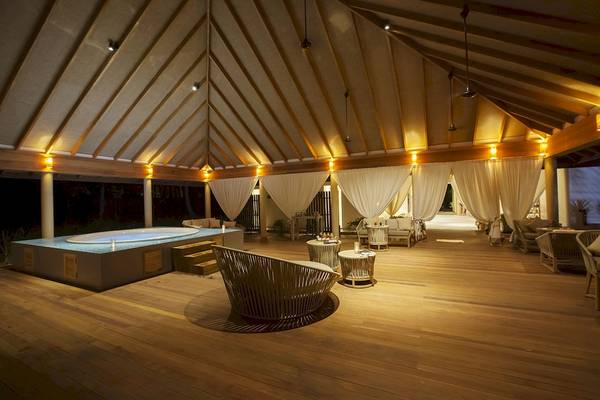 Innenbereich des Reethi Faru Resorts mit einem Whirlpool, umgeben von Holzmöbeln und weißen Vorhängen.