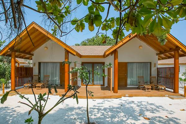 Außenansicht des Reethi Faru Resorts mit hölzernen Bungalows und Verandas, umgeben von weißem Sand und Bäumen.