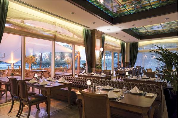 Innenansicht des Restaurants im Voyage Belek Golf & Spa Hotel mit gedeckten Tischen und grünen Vorhängen.