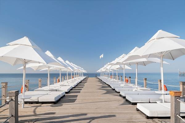 Mehrere weiße Sonnenschirme und Liegen auf einem Pier am Meer, im Hintergrund ein blauer Himmel und eine Flagge.