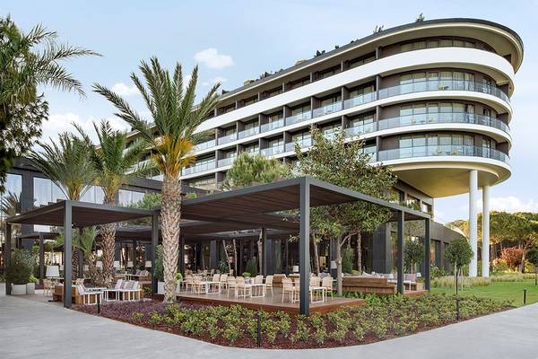 Außenbereich des Voyage Belek Golf & Spa Hotels mit moderner Architektur, Terrassenmöbeln und Palmen.