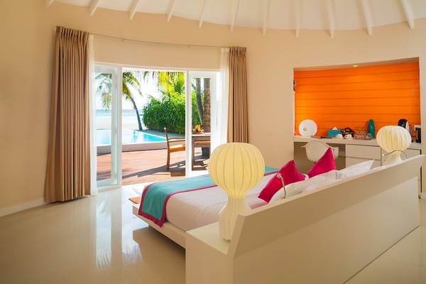Innenansicht eines Zimmers im Sun Siyam Vilu Reef Hotel mit Bett, Lampen und Poolsicht.