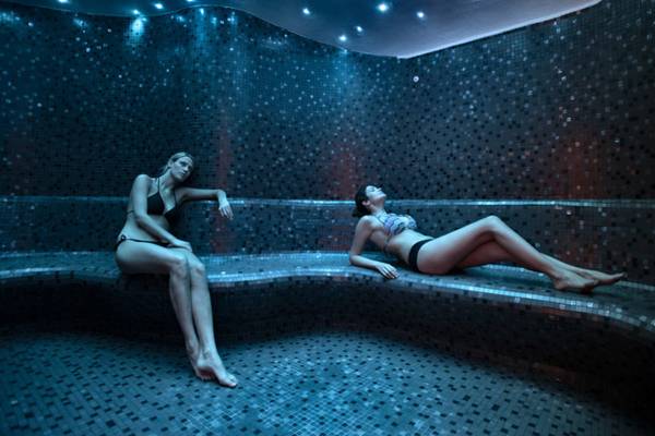 Zwei Frauen entspannen in einer gefliesten Sauna mit blauer Beleuchtung.