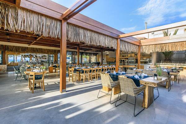 Restaurantbereich des Stella Village Hotel & Bungalows mit hölzernen Tischen und Stühlen unter einer Strohdach-Überdachung.