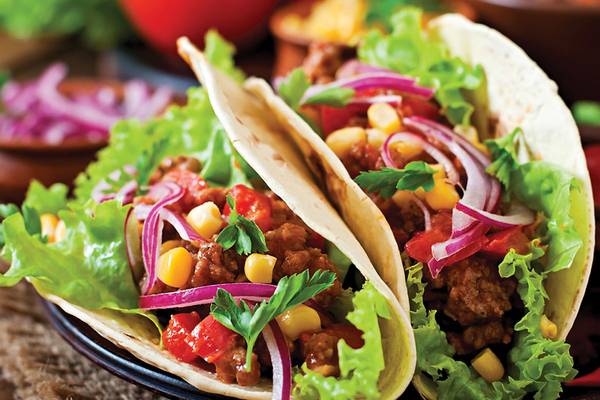 Zwei Tacos mit Hackfleisch, Salat, Tomaten, Mais und roten Zwiebeln auf einem schwarzen Teller.