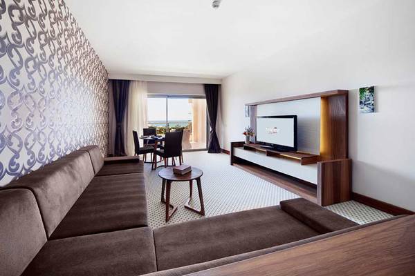 Wohnzimmer im Sueno Hotels Beach Side mit einem Sofa, einem Tisch, einem Fernseher und einem Esstisch mit Stühlen.