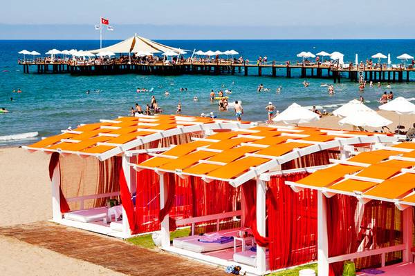 Strandszenerie mit Sueno Hotels Beach Side, mehreren orangefarbenen Strandhütten und vielen Gästen.