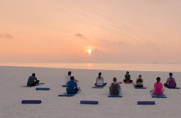 Gruppe von Menschen praktiziert Yoga auf dem weißen Sandstrand des Meeru Island Resort & Spa bei Sonnenuntergang