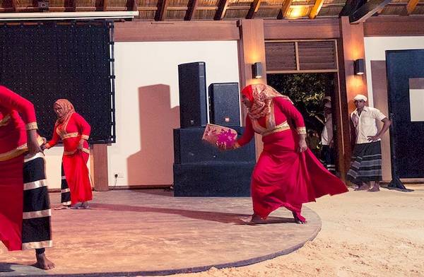 Tanzgruppe im Meeru Island Resort & Spa, alle in roten Kleidern, vor einer Bühne mit Lautsprechern und einem Mann, der zuschaut.