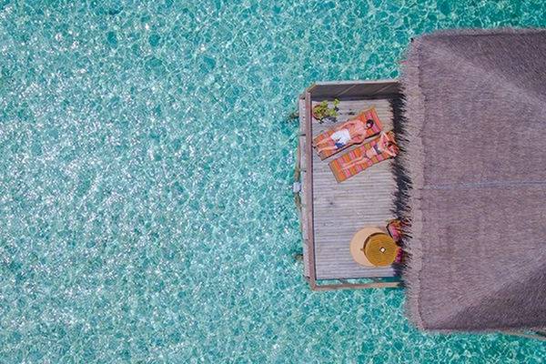 Luftaufnahme des Meeru Insel Resorts & Spa Wasserbungalow mit zwei Personen, die auf Liegen entspannen.