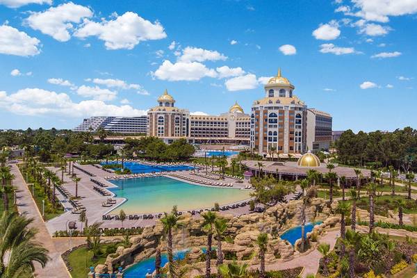 Hotelansicht des Delphin BE Grand Resorts mit Schwimmbad und tropischem Garten unter einem blauen Himmel.