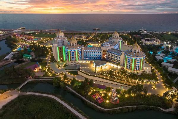Luftaufnahme des Delphin BE Grand Resort mit bunten Lichtern und Blick auf das Meer bei Sonnenuntergang.
