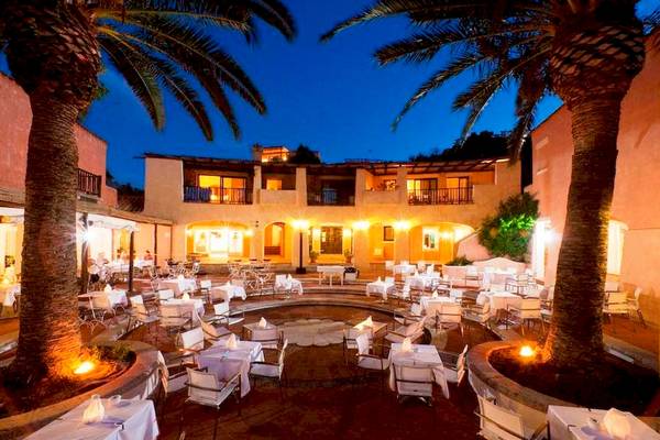 Abendlicher Außenbereich des Bagaglino I Giardini di Porto Cervo Hotels mit runden Tischen und Palmen.