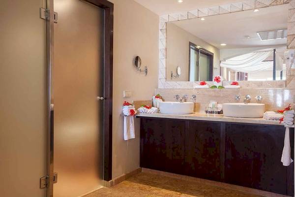 Badezimmer mit Doppelwaschbecken im Majestic Elegance Punta Cana Hotel, dekoriert mit Blumen und Handtüchern.