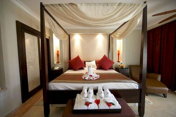 Hotelzimmer im Majestic Elegance Punta Cana mit Himmelbett, roten Kissen und weißen Hausschuhen auf einem Tablett.