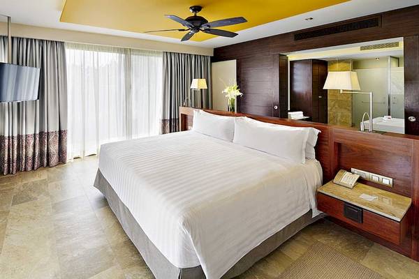 Ein modernes Hotelzimmer im Barcelo Maya Grand Resort mit einem großen Bett, Fernseher, und Badezimmerspiegel.