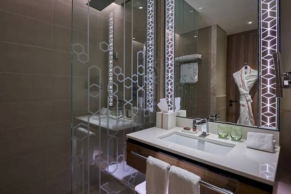 Badezimmer im Mysk Al Mouj Hotel mit gefliester Wand, Glasdusche und beleuchtetem Spiegel.