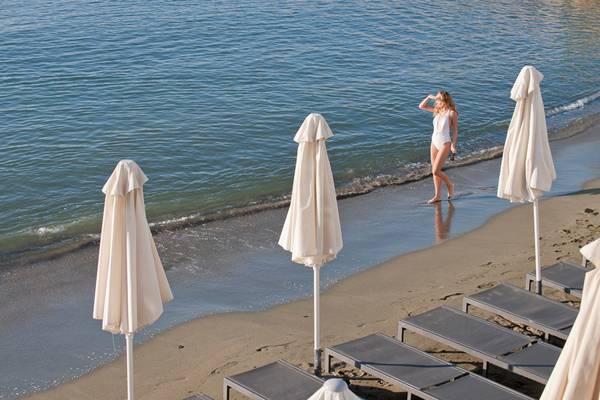 Frau im Badeanzug am Strand des Royal Apollonia Beach Hotels, vor einem blauen Ozean und weißen Sonnenschirmen.