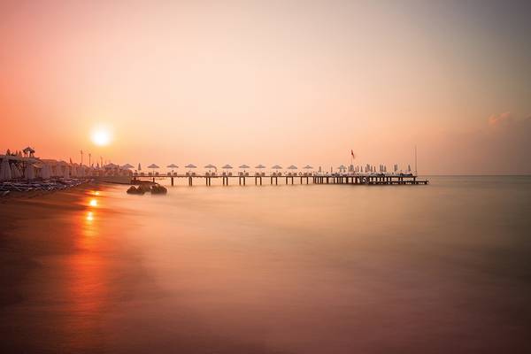 Ein Strand mit einem Pier und dem Sonnenuntergang im Hintergrund, reflektiert auf dem Wasser.