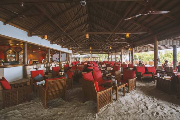 Loungebereich des Reethi Beach Resorts mit roten Stühlen und hölzernen Tischen auf Sand.