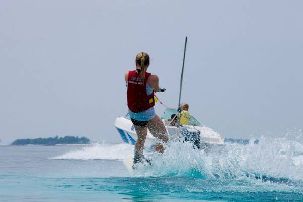 Frau im roten Vest auf Wasserskis im Meer, ein Boot im Hintergrund