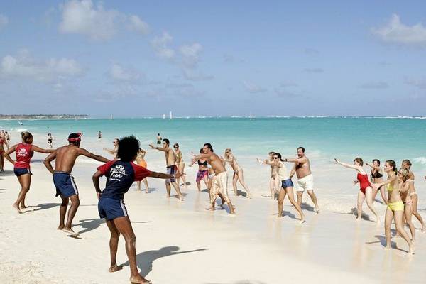 Gruppe von Menschen, die an einem Strand unter blauem Himmel tanzen, einige tragen Badebekleidung, im Hintergrund das Meer