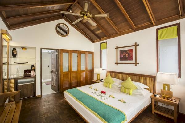 Innenansicht eines Zimmers im Reethi Beach Resort mit einem Bett, Deckenventilator und Nachttischen.