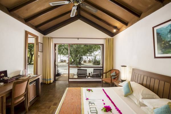 Innenansicht eines Zimmers im Reethi Beach Resort mit einem Bett, einem Schreibtisch, einem Ventilator und einem Fenster, das zu einer Terrasse mit Blick auf den Strand führt.