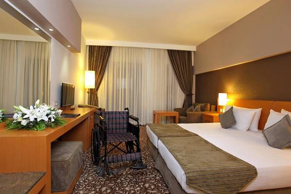 Hotelzimmer mit zwei Betten, Schreibtisch, Rollstuhl und Blumenstrauß.