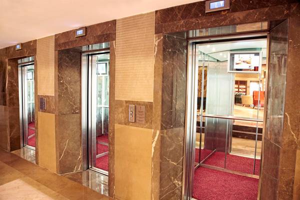 Der Innenbereich eines Hotels zeigt drei Aufzüge mit Marmorwänden und roten Teppichböden.