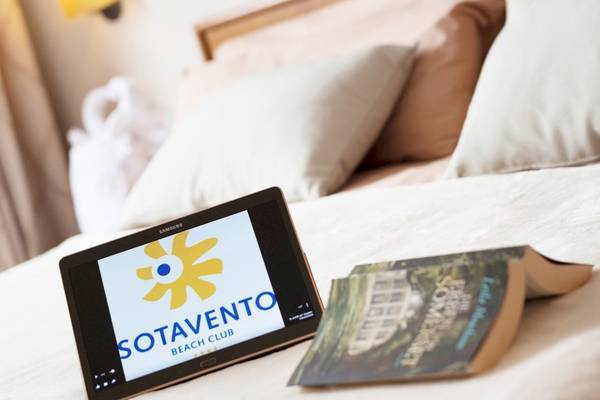 Ein Tablet mit dem Logo von Sotavento Beach Club auf einem Bett neben einem Buch.