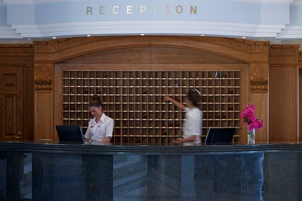 Rezeptionsbereich des Mitsis Summer Palace Beach Hotels mit zwei Angestellten und einer Frau, die die Schlüssel abholt.