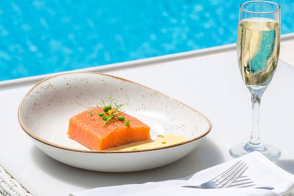 Ein Teller mit Lachs in einer Porzellanschale, neben einem Glas Champagner, vor einem Pool.
