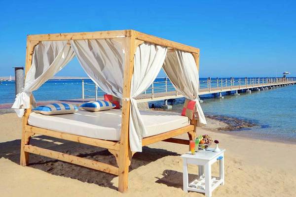 Ein Bett am Strand des Pickalbatros White Beach Resorts mit einem weißen Vorhang und einer kleinen Tisch daneben.