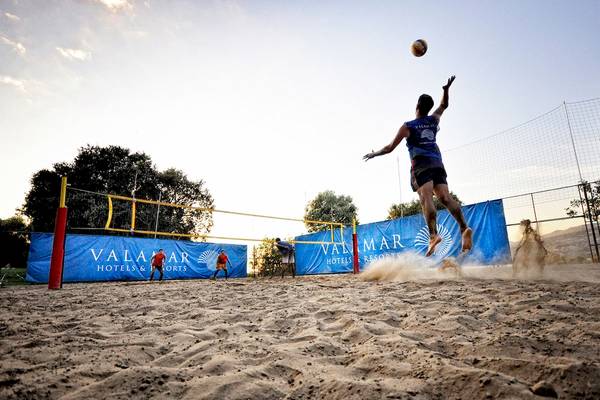 Mann springt und schlägt Volleyball auf Sandplatz bei Sonnenuntergang, VALMAR Hotels & Resorts im Hintergrund.