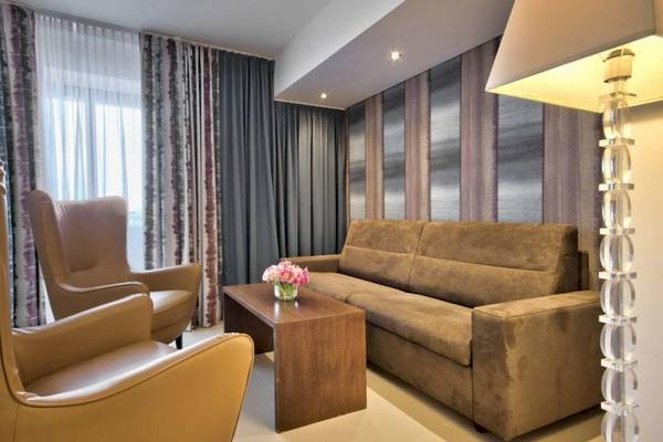 Innenansicht des AX The Palace Hotels mit einem Wohnzimmer, Sofa, zwei Ledersesseln, Tisch, Blumenvase und einer Stehlampe.