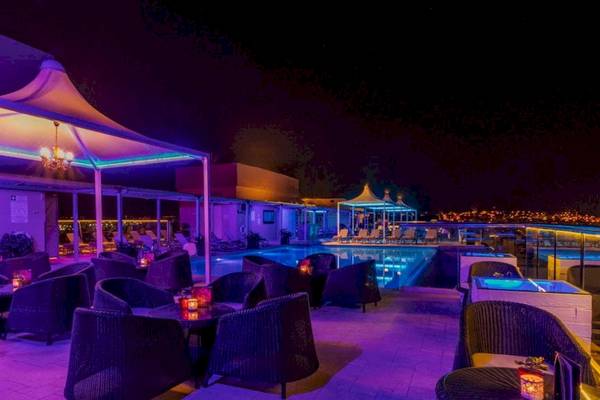 Die Pool-Lounge-Area des AX The Palace Hotels bei Nacht mit lila Beleuchtung und runden Tischen.