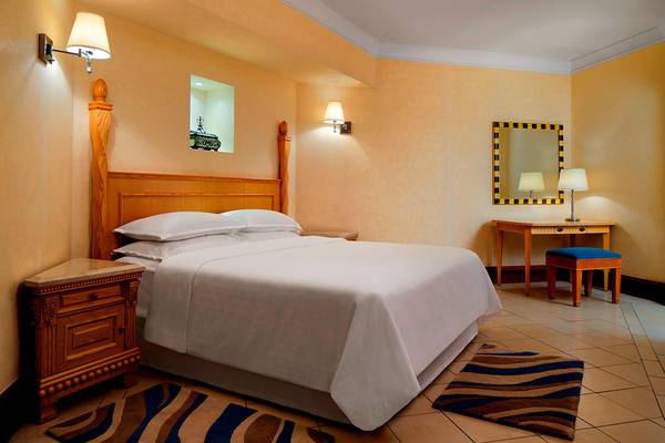 Ein Schlafzimmer im Sheraton Jumeirah Beach Resort mit einem großen Bett, weißen Laken, Holzmöbeln und dekorativen Elementen.