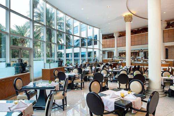 Innenansicht des Restaurants im Sheraton Jumeirah Beach Resort mit runden Tischen und Stühlen, großen Fenstern und Palmen im Hintergrund.