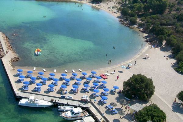 Luftaufnahme des Hotel Ollastu Resorts mit Strand, blauen Sonnenschirmen und Booten im Wasser.