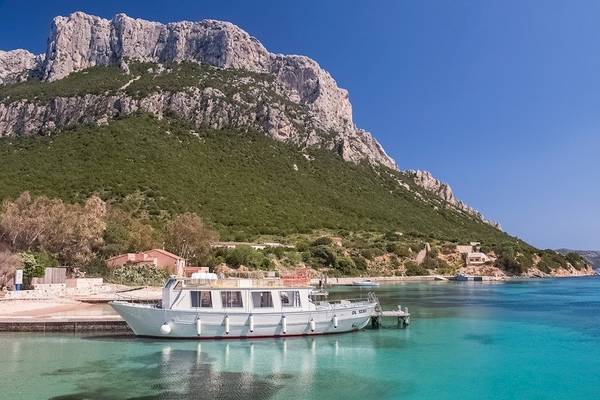 Ein weißer Boot am Kai neben dem Hotel Ollastu Resort mit einer felsigen Berg im Hintergrund.