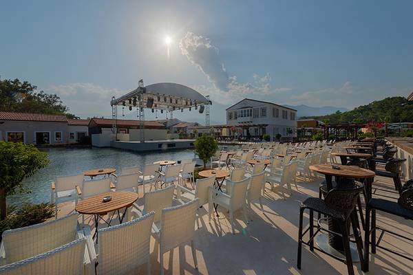 Außenterrasse des Rixos Premium Tekirova Hotels mit vielen Tischen und Stühlen, neben einem Wassergraben und einer Bühne.